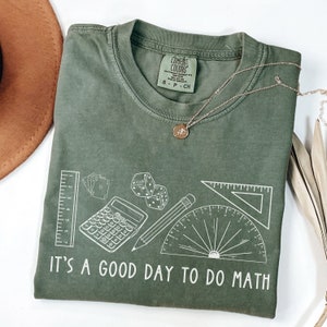 May include: A green t-shirt with a white graphic of math-related items, including a ruler, calculator, pencil, protractor, dice, and playing cards. The text "IT'S A GOOD DAY TO DO MATH" is printed below the graphic.