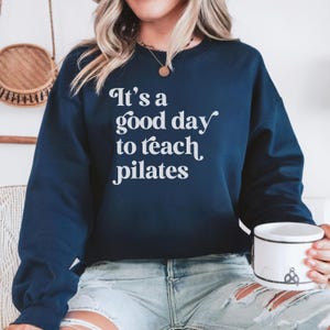 May include: Navy blue sweatshirt with the text "It's a good day to teach pilates" in white script. The sweatshirt is a crew neck style and is made of a soft material. The image shows a person wearing the sweatshirt.