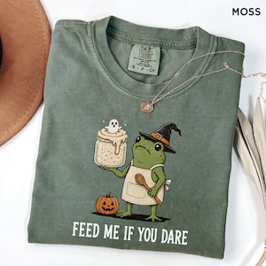 Könnte beinhalten: Ein moosgrünes T-Shirt mit einer Grafik eines grünen Frosches, der einen Hexenhut und eine Schürze trägt und ein Glas mit einem Geist sowie einen Holzlöffel hält. Ein kleiner orangefarbener Kürbis steht daneben. Der Text lautet: "FEED ME IF YOU DARE."