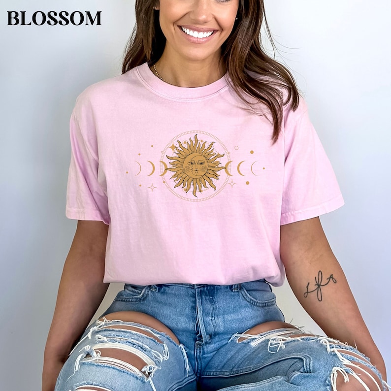 May include: A pink t-shirt with a gold sun and moon graphic. The sun has a face and is surrounded by stars and crescent moons.
