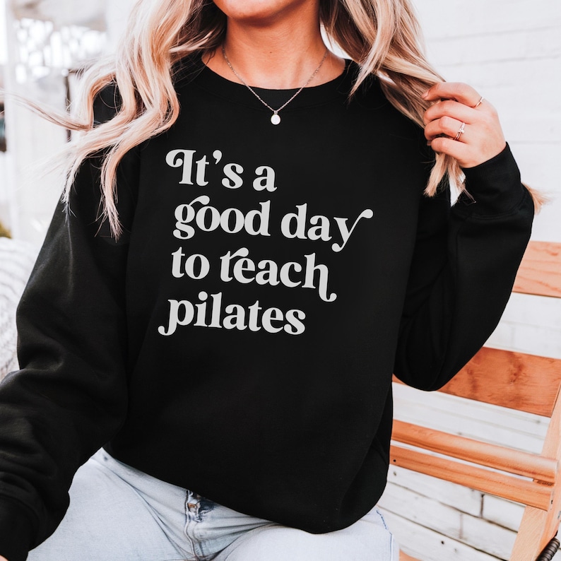May include: Black sweatshirt with the white text "It's a good day to teach pilates". The sweatshirt is a crew neck style. The image shows a person wearing the sweatshirt.