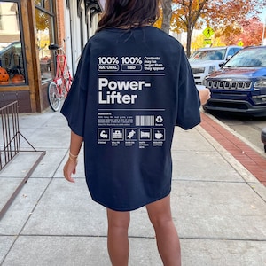 May include: Black oversized t-shirt with a white graphic design that says "100% Natural 100% SBD Power-Lifter" and includes a list of ingredients: "Strong, Will PR, Bund on Candy, Nap Between Sets, Requires Caffeine".