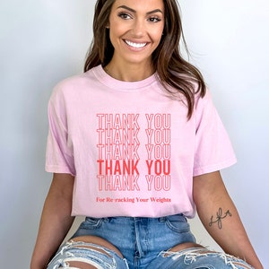 Peut inclure: T-shirt rose avec le texte "THANK YOU" r&eacute;p&eacute;t&eacute; cinq fois en rouge. Le texte "For Re-racking Your Weights" est imprim&eacute; sous le texte r&eacute;p&eacute;t&eacute;.