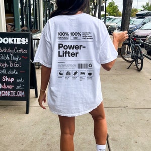 May include: White t-shirt with a black graphic on the back that says "100% 100% NATURAL SBD Power-Lifter". The graphic also includes icons for strong, will power, suns on candy, naps between sets, and requires caffeine.