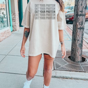 May include: A beige oversized t-shirt with the text "EAT YOUR PROTEIN" repeated five times in black.