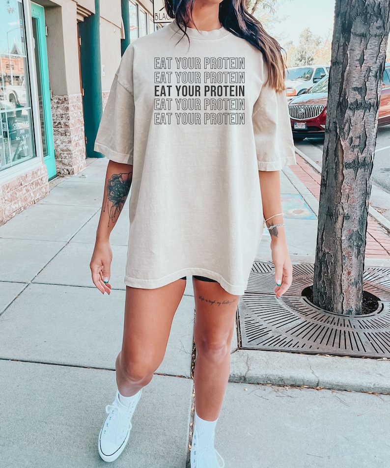 May include: A beige oversized t-shirt with the text "EAT YOUR PROTEIN" repeated five times in black.
