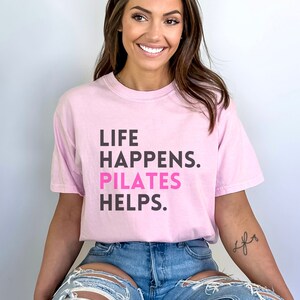 May include: A pink t-shirt with the text "LIFE HAPPENS. PILATES HELPS." in black and pink lettering. The model is wearing the shirt with blue jeans.