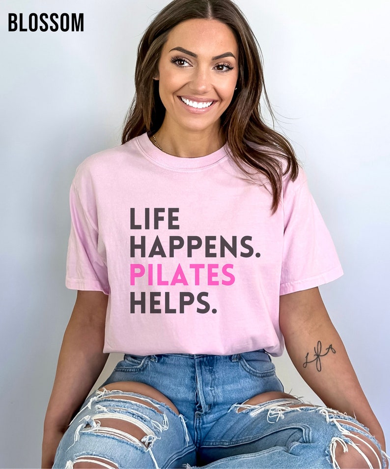 May include: A pink t-shirt with the text "LIFE HAPPENS. PILATES HELPS." in black and pink lettering. The model is wearing the shirt with blue jeans.
