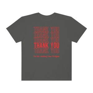 Peut inclure: Un t-shirt gris fonc&eacute; avec le texte "THANK YOU" r&eacute;p&eacute;t&eacute; six fois en rouge. Le texte est empil&eacute; verticalement et le bas du t-shirt indique "For Re-racking Your Weights".
