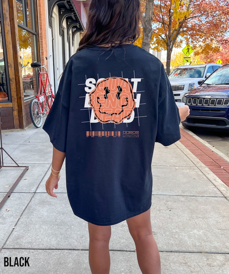 May include: Black oversized t-shirt with a graphic of a smiley face and the words "SQUAT BENCH DEAD" printed on the back. The graphic is in orange and white.