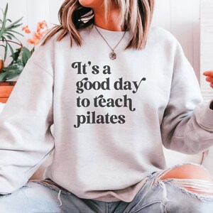 May include: A light gray sweatshirt with the text "It's a good day to teach pilates" in black script. The sweatshirt is a crew neck style and appears to be made of a soft material. The overall theme is fitness and pilates.