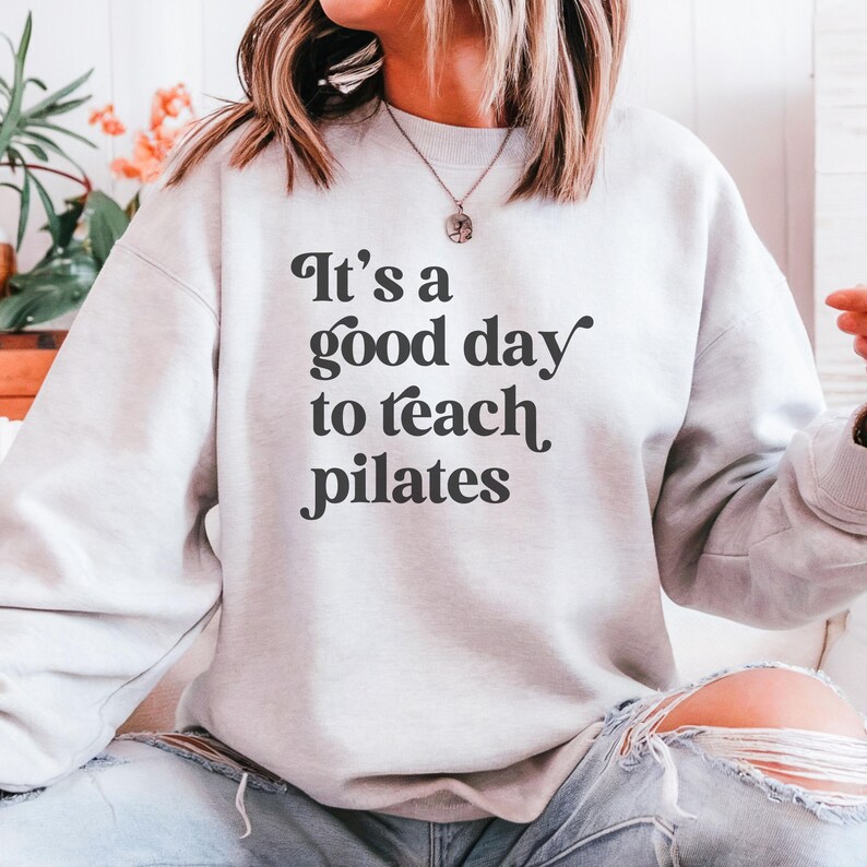May include: A light gray sweatshirt with the text "It's a good day to teach pilates" in black script. The sweatshirt is a crew neck style and appears to be made of a soft material. The overall theme is fitness and pilates.