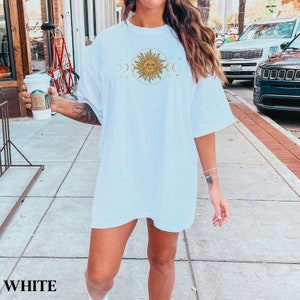 May include: White oversized t-shirt with a graphic of a sun with a face and moons on either side. The text "WHITE" is visible at the bottom of the image.