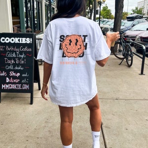 May include: White t-shirt featuring a graphic of a smiley face with the words "SQUAT BENCH DEAD" surrounding it. The text is in a black grid pattern.