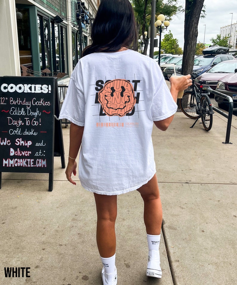 May include: White t-shirt featuring a graphic of a smiley face with the words "SQUAT BENCH DEAD" surrounding it. The text is in a black grid pattern.