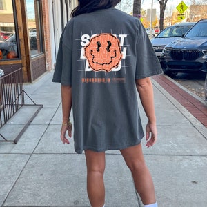 May include: A charcoal grey t-shirt with a graphic print of a smiley face with the words "SQUAT BENCH DEAD" printed inside the face. The graphic is overlaid on a grid pattern.