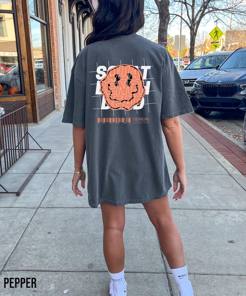 May include: A charcoal grey t-shirt with a graphic print of a smiley face with the words "SQUAT BENCH DEAD" printed inside the face. The graphic is overlaid on a grid pattern.