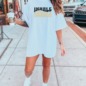 Op de afbeelding: Wit oversized T-shirt met de tekst "INHALE EXHALE" in een retro lettertype. De tekst is in zwart en geel.