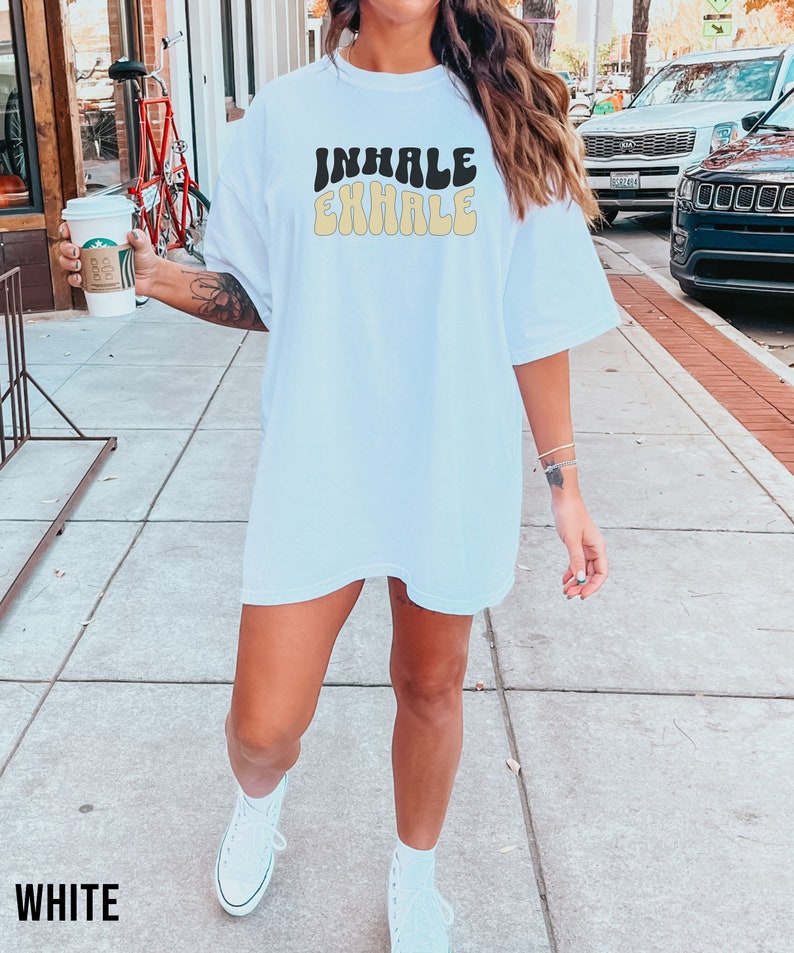 Op de afbeelding: Wit oversized T-shirt met de tekst "INHALE EXHALE" in een retro lettertype. De tekst is in zwart en geel.