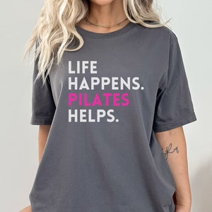 May include: Gray short-sleeved t-shirt with the text "LIFE HAPPENS. PILATES HELPS." in white and pink.