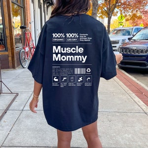 Puede incluir: Camiseta negra de gran tama&ntilde;o con un estampado gr&aacute;fico de una etiqueta nutricional para "Muscle Mommy". La etiqueta incluye el texto "100% Org&aacute;nico 100% D&iacute;a de Piernas" y "El contenido puede ser m&aacute;s fuerte de lo que parece".