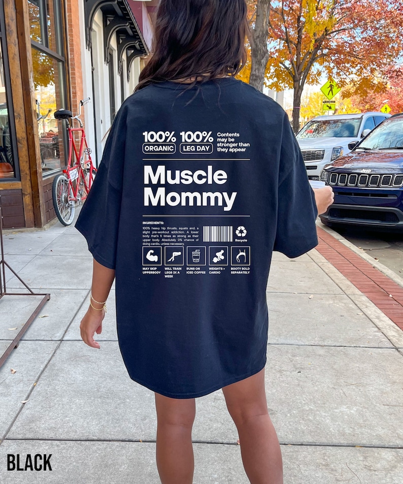 Puede incluir: Camiseta negra de gran tama&ntilde;o con un estampado gr&aacute;fico de una etiqueta nutricional para "Muscle Mommy". La etiqueta incluye el texto "100% Org&aacute;nico 100% D&iacute;a de Piernas" y "El contenido puede ser m&aacute;s fuerte de lo que parece".