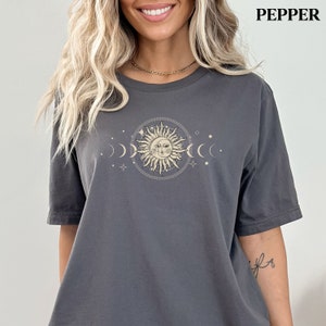 May include: A gray t-shirt with a graphic of a sun and moon with stars. The sun has a face and is surrounded by a circle of smaller circles. The moon is in a crescent shape and is on either side of the sun.