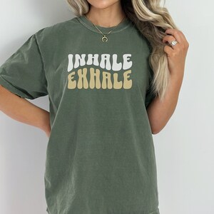Op de afbeelding: Een groen T-shirt met de woorden "INHALE EXHALE" in een retro-lettertype. De woorden zijn wit en geel.