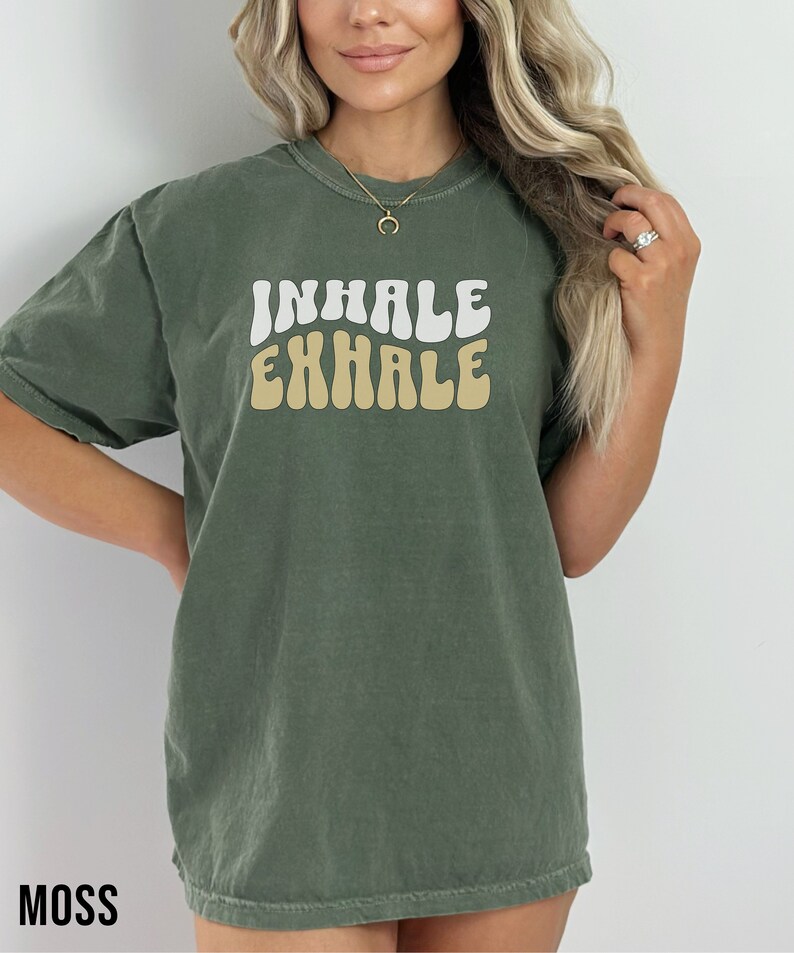 Op de afbeelding: Een groen T-shirt met de woorden "INHALE EXHALE" in een retro-lettertype. De woorden zijn wit en geel.