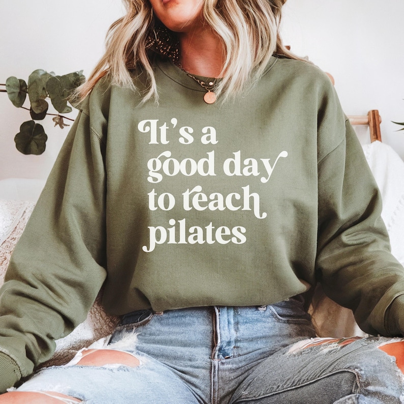 May include: Olive green sweatshirt with the text "It's a good day to teach pilates" in white script. The sweatshirt is a crew neck style and is made of a soft material. The image shows the sweatshirt being worn.