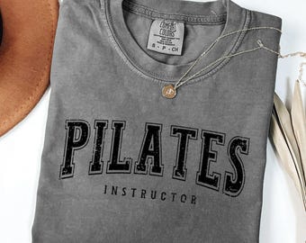 Könnte beinhalten: Ein graues T-Shirt mit dem Text "PILATES INSTRUCTOR" in schwarzen Buchstaben. Das Shirt hat einen Used-Look und ist aus einem weichen, bequemen Stoff.