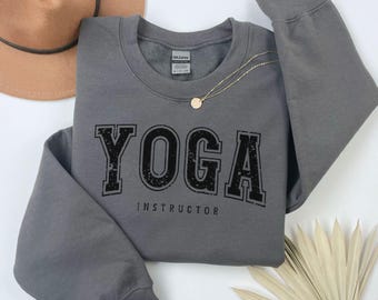 May include: Charcoal grey sweatshirt with the text "YOGA! INSTRUCTOR" in black. The sweatshirt has a crew neck and long sleeves.