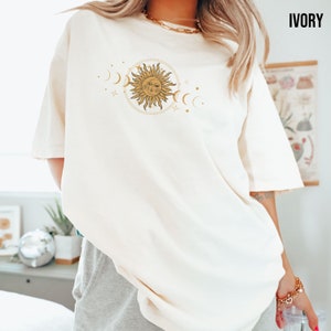 May include: A white oversized t-shirt with a gold sun and moon graphic. The graphic features a sun with a face and rays, surrounded by crescent moons and stars.