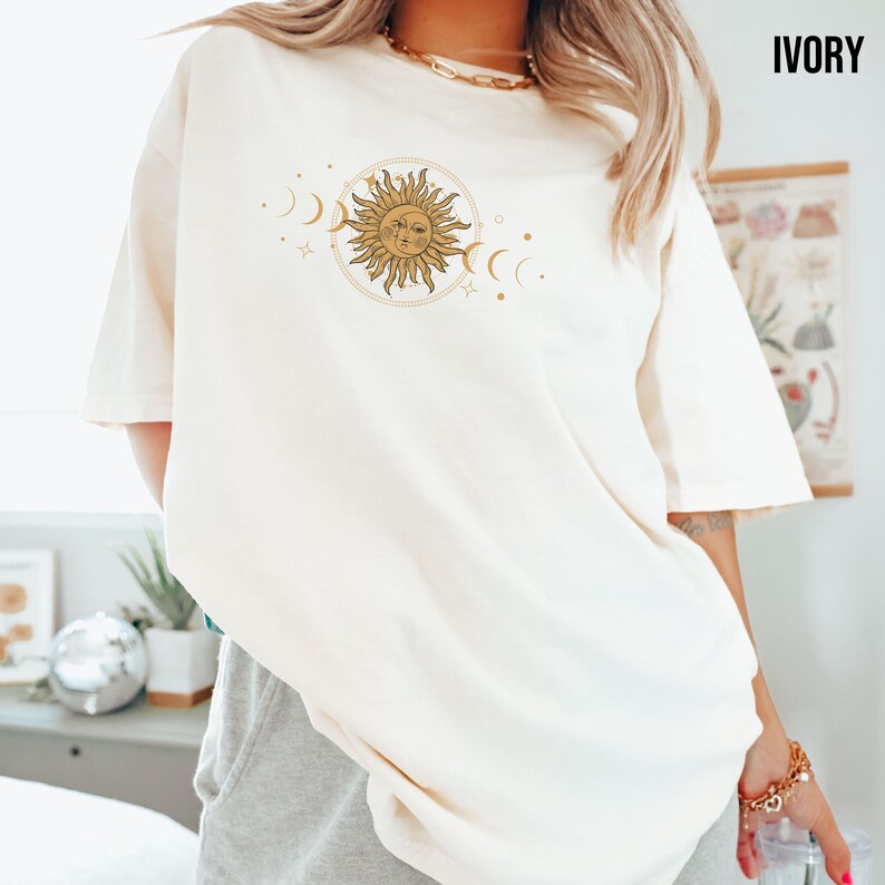May include: A white oversized t-shirt with a gold sun and moon graphic. The graphic features a sun with a face and rays, surrounded by crescent moons and stars.