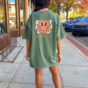 May include: An oversized green t-shirt with a graphic of a smiling face and the text "SOUT BEING DEAD" printed on the back.