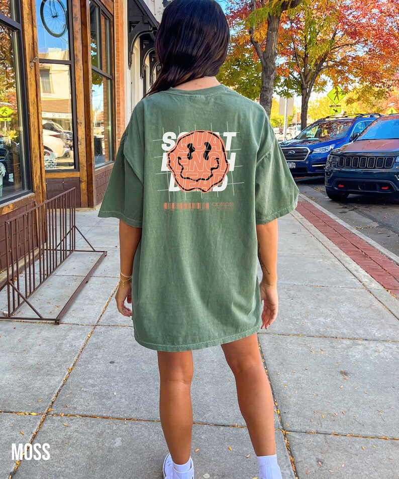 May include: An oversized green t-shirt with a graphic of a smiling face and the text "SOUT BEING DEAD" printed on the back.
