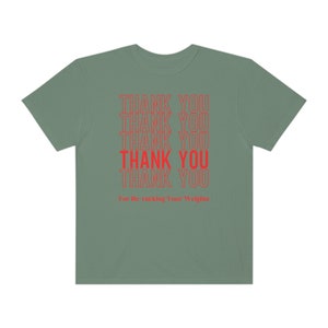 Peut inclure: Un t-shirt vert sauge avec le texte "THANK YOU" r&eacute;p&eacute;t&eacute; six fois en rouge. Le texte est empil&eacute; verticalement et la ligne du bas indique "For Re-racking Your Weights".