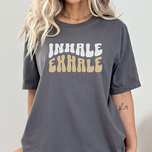 Op de afbeelding: Een antracietgrijs T-shirt met een witte en beige retro-stijl grafische afbeelding met de tekst "INHALE EXHALE".
