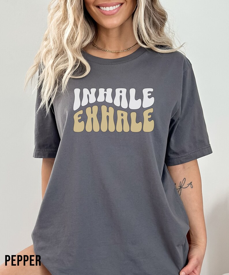 Op de afbeelding: Een antracietgrijs T-shirt met een witte en beige retro-stijl grafische afbeelding met de tekst "INHALE EXHALE".