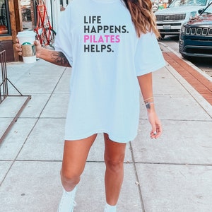 May include: White oversized t-shirt with the text "LIFE HAPPENS. PILATES HELPS." in black and pink lettering.