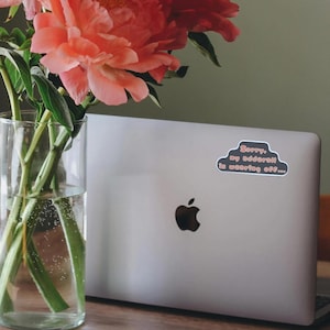 May include: A silver laptop with a black Apple logo and a sticker that reads "Sorry, my adderall is wearing off..." is next to a glass vase with pink flowers.
