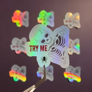 May include: A holographic sticker with a skeleton design and the text "TRY ME". The sticker also features a small spider graphic.