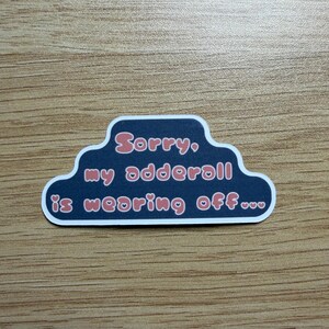 May include: A sticker with a dark blue cloud-shaped border and white trim. The text "Sorry, my adderall is wearing off..." is written in pink, stylized font. The sticker is on a wooden surface.