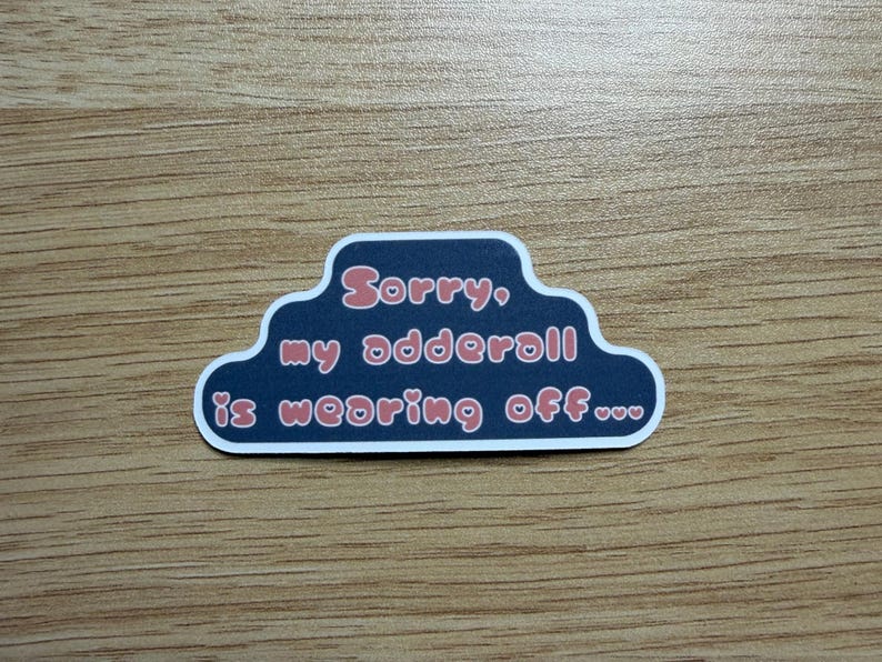 May include: A sticker with a dark blue cloud-shaped border and white trim. The text "Sorry, my adderall is wearing off..." is written in pink, stylized font. The sticker is on a wooden surface.