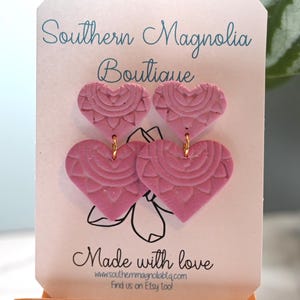 May include: Pink heart-shaped earrings with a detailed mandala design, displayed on a white card with the text "Southern Magnolia Boutique" and "Made with love". The earrings hang from gold-colored hooks.
