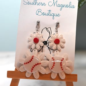 Könnte beinhalten: Weiße Gänseblümchen-Ohrringe mit roten Zentren und Baseball-Akzenten. Die Ohrringe werden auf einer kleinen Holzstaffelei mit dem Text "Southern Magnolia Boutique" auf der Karte präsentiert.