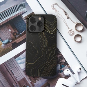 May include: A black iPhone case with a gold topographic design. The case is lying on a white surface with other items, including a magazine, sunglasses, and jewelry.