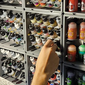 May include: A wall-mounted storage system with multiple shelves filled with small bottles of paint. The bottles are organized by color and are labeled with their names. A person's hand is reaching into one of the shelves to grab a bottle of paint.