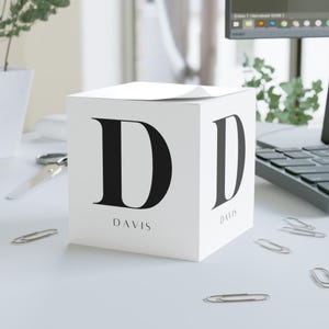 May include: White cube-shaped notepad with a large black letter "D" on two sides. The name "DAVIS" is printed below the letter. Several silver paper clips are scattered around the notepad on a white surface.