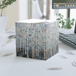 May include: A square notepad featuring a birch tree forest design with butterflies and a small bird against a blue background. The notepad is sitting on a white surface with paper clips scattered around.